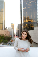 Beautiful young woman sitting on a bridge across the boulevard in urban scenery, downtown, at sunset, smiling at camera.