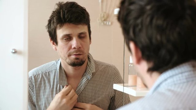 Tired man who has just woken up looks at his reflection in the mirror and sees his scruffy appearance. Buttoning a plaid shirt.