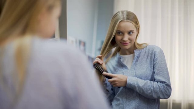 Smiling beautiful female combing long blond hair near mirror, haircare treatment