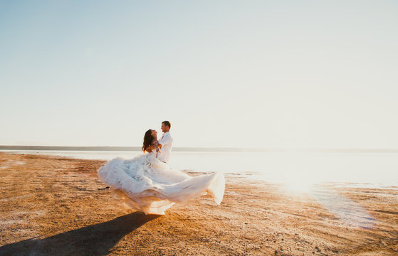 Wedding couple is spinning on the sea beach. Sunny summer photo. Bride with hair down in off shoulder dress with train. Ocean romantic ceremony. Seaside love story. Sand, water and horizon. Honeymoon.