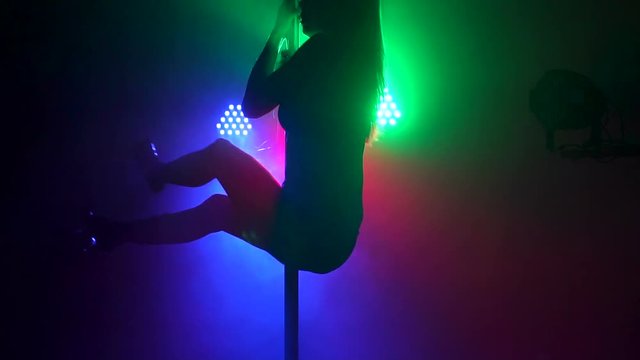 A flexible dancer performs a complex exercise on a pylon in the dark with the light of green, red and blue projectors. Pole dance. Slow motion. Silhouette.