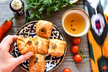 woman's hand hold samosa and delicious Uzbek pieces of samosas with cherry tomatoes, Chile on the plate with Uzbek ornament on a wooden background in rustic style for breakfast, uzbek utensils - obrazy, fototapety, plakaty