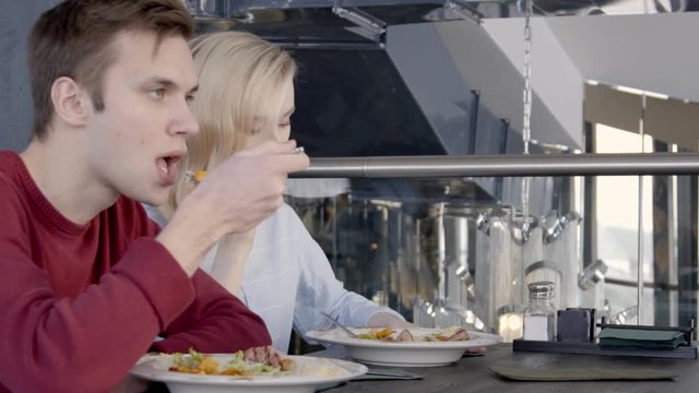 Two colleagues are having luch in restaurant together, slow motion. Blond woman in blue shirt is talking on her brand-new smartphone and man in red jumper are sitting near at the wooden table and