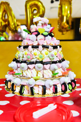 A cake made with sugary and chewy candies, chupa chups on a table.
Red tablecloth with white balls. Blurred background with inflated letters