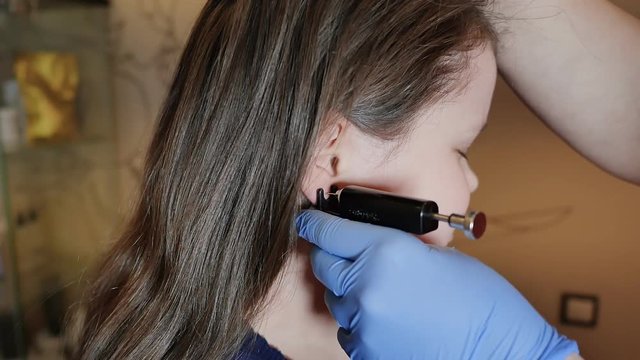 Adorable little girl having ear piercing process with special equipment in beauty center by medical worker