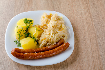 Roasted sausages with steamed cabbage (sauerkraut) and boiled potatoes in white plate.