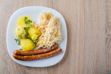 Roasted sausages with steamed cabbage (sauerkraut) and boiled potatoes in white plate.