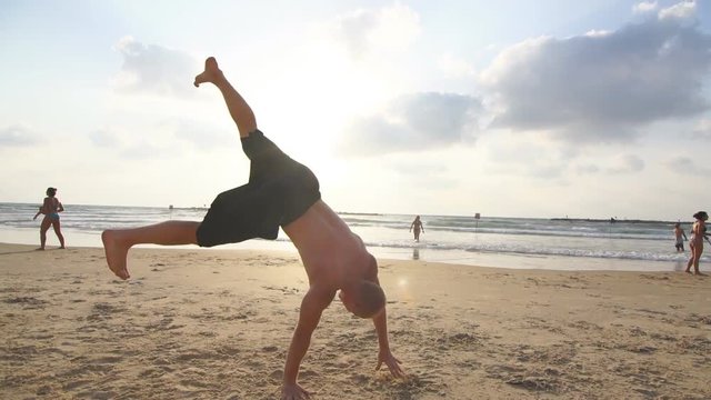 Young man doing parkour tricks on sea beach at sunset. Freerunner guy showing various jumps while running on ocean shore at sundown. Acrobatic stunts and somersaulting on sunny summer day. Slow motion