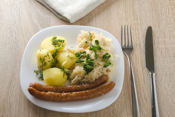 Roasted sausages with steamed cabbage (sauerkraut) and boiled potatoes in white plate.