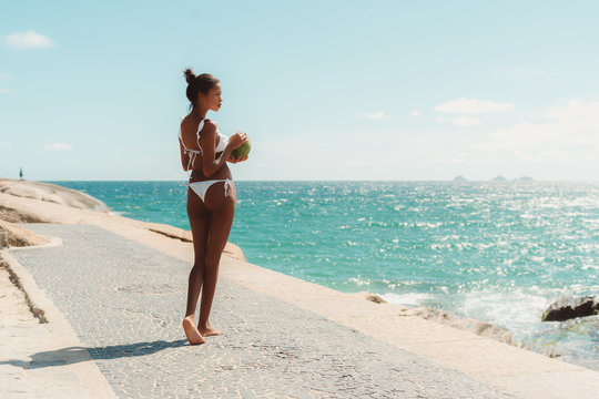 Rear view of sexy Brazilian girl with slim model body standing on paving stone near ocean beach, half-turned to the camera and pensively looking aside, holding in hands fresh coconut with straw