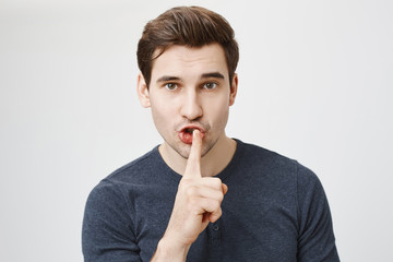 Close-up portrait of handsome athletic guy showing shh gesture with index finger near mouth, expressing concern while standing against gray background. I think we should keep it a secret