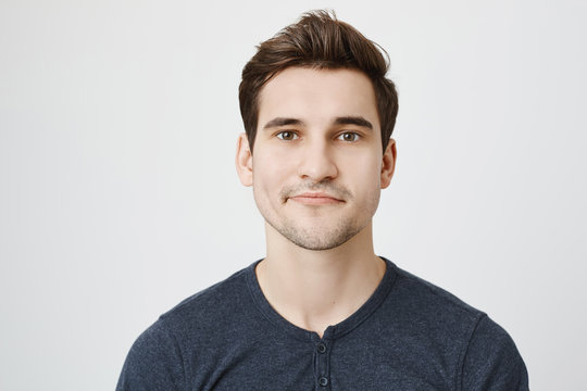 Close-up portrait of nice european guy with stylish haircut and curvy smile, standing against gray background. Student came to his teacher to pass exam and feels awkward for not attending her classes