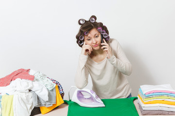 Distressed housewife in light clothes with curlers on hair talking on mobile phone, ironing family clothing on ironing board with iron. Woman isolated on white background. Copy space for advertising.