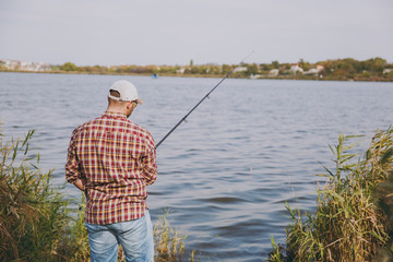 Back view Young unshaven man with fishing rod in checkered shirt, cap and sunglasses casts bait and...