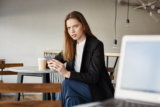 Woman is insulter with offensive words of stranger sitting with laptop. Portrait of annoyed attractive woman in cafe, holding smartphone and drinking coffee, frowning at person who tells rude things