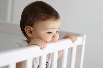 Portrait of baby staying in his baby cot looking sadly outside with frowned brows