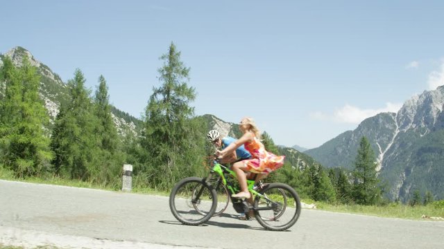 Struggling road cyclist waves and comments on being easily overtaken by girl on innovative electric bike. Blonde city woman in sundress pedals past pro mountain bike rider on sunny day in the summer.