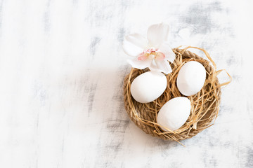 Three white Easter eggs lying on a tray made of twine, near one orchid. White wooden vintage background. Copy space