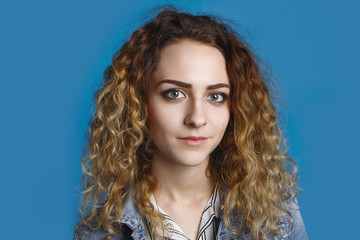 Portrait of beautiful student girl with clean perfect skin and wavy light hair posing in studio, dressed in denim jacket against blank blue wall background with copy space for your advertising content