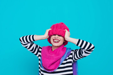 Close up face portrait of toothy smiling young woman wearing knitted pink hat and scarf. A happy smiling woman on a turquoise background in the studio. 
