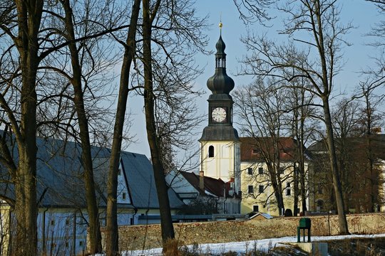 Winter picture of church tower with clock / premises of Zdar nad Sazavou castle, trees, wall, white building of museum, bright blue sky
