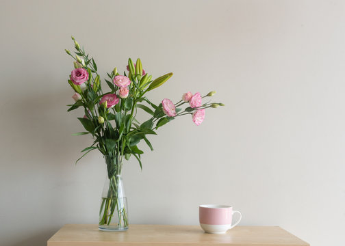Pink lisianthus flowers in glass vase with mug on wooden shelf against beige wall