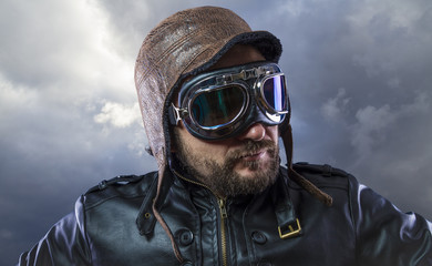 Old plane pilot on background of storm clouds with expressive face. glasses and old hat with black leather jacket