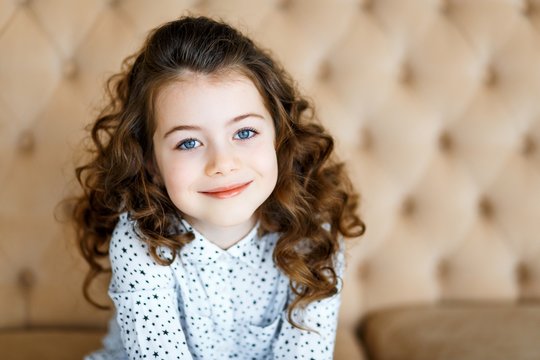 A photo of a beautiful little girl with blue eyes and brunette curly hair in studio sitting on the sofa and smoling