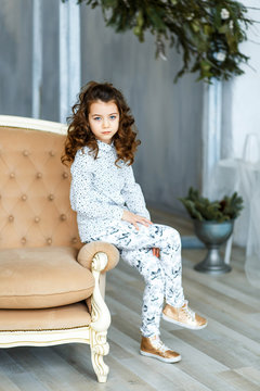 A photo of a beautiful little girl with blue eyes and brunette curly hair in studio sitting on the sofa and smoling