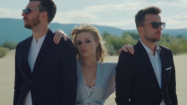 Blonde is standing between two boyfriends in a desert. Strange relationships of woman with two man, abstract shot on a beach