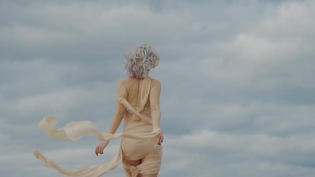 Elegant woman is wearing eccentric suit with waving fabric strips is going out from camera. Back view of her amazing slim body, picturesque clouds in background