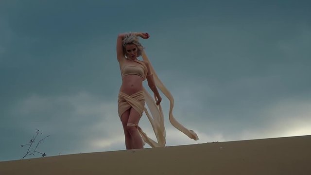 Sexy young blonde is posing outdoors on a sand in background of rainy sky. She is touching scraps of her dress swaying by wind, abstract and fashionable shot