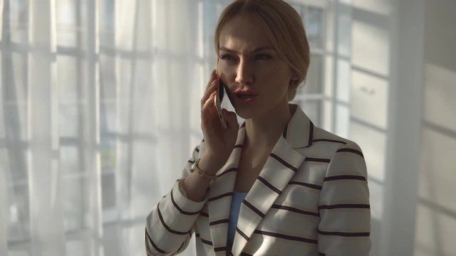 Young blond woman talking on the phone next to a large window.