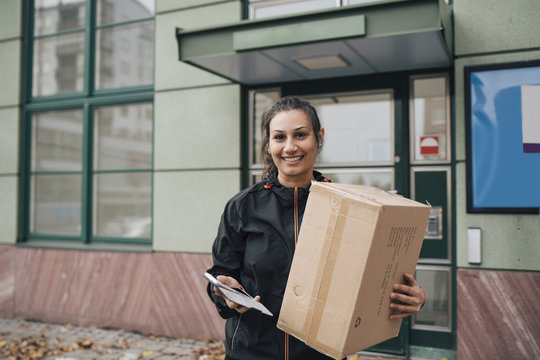 Portrait of smiling worker carrying box and holding phone while standing against building in city