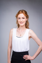 Fototapeta premium portrait vertical up to the waist young, beautiful business woman, student with lred, curly hair and freckles on gray background. SHe dressed in a white blouse with short sleeves about open shoulders