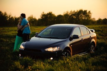 A loving couple watching beautiful bright romantic sunset after journey, standing leaning against blue sport car. The fields around them. The young man shows to his girlfriend evening sun
