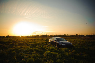 Fototapeta premium Blue sport car with headlights in a field in bright sunset light at summer evening