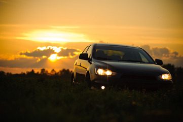 Fototapeta premium Blue sport car with headlights in a field in bright sunset light at summer evening
