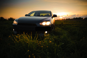 Fototapeta na wymiar Blue sport car with headlights in a field in bright sunset light at summer evening