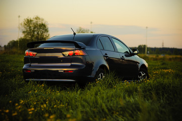 Fototapeta premium Blue sport car with headlights in a field in bright sunset light at summer evening