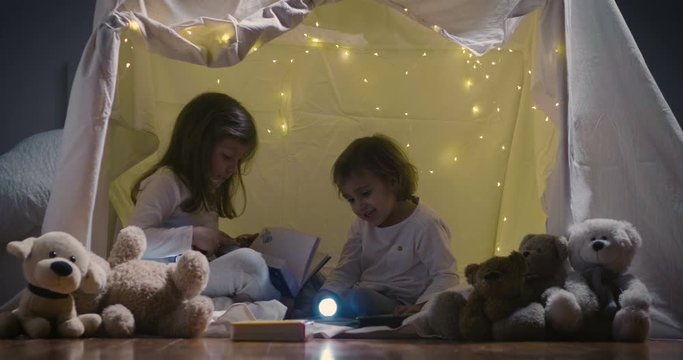 Two little girls play at home in the evening to build a camping tent to read books with a flashlight and sleep inside. Concept of: game, magic, creativity, alarm systems.