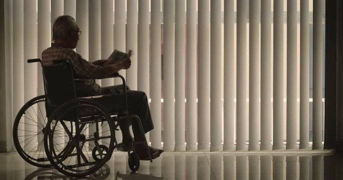 Lonely senior man reading a newspaper while sitting on wheelchair near the window with curtain at home. Shot in 4k resolution
