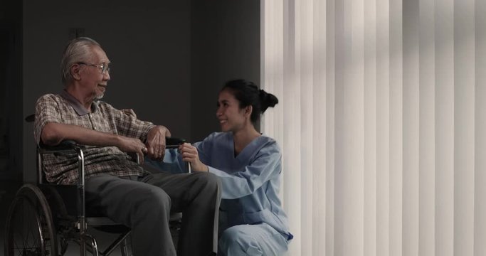 Elderly man sitting on wheelchair while chatting with his nurse and pushed by her near the window in the hospital. Shot in 4k resolution