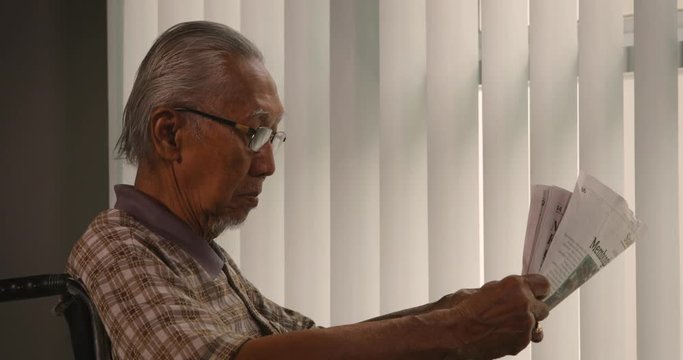 Elderly man with grizzled hair wearing glasses and reading a newspaper while sitting on wheelchair near the window at home. Shot in 4k resolution