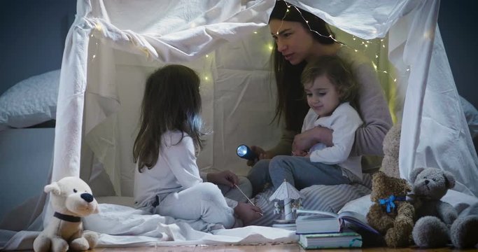 Two little girls play at home in the evening to build a camping tent to read books with a flashlight and sleep inside. Concept of: game, magic, creativity, alarm systems.