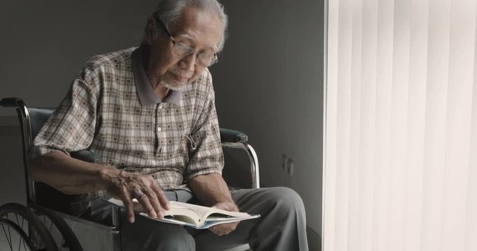Elderly Asian man reading a book alone while sitting on the wheelchair near the window at home. Shot in 4k resolution