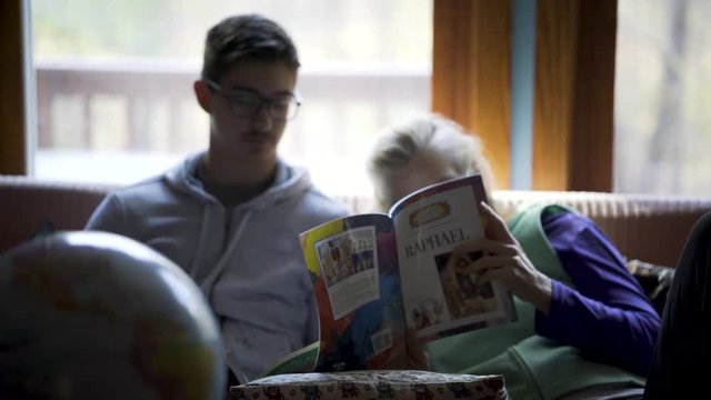 Camera pulling away on a mother and teenage son doing homeschool lessons in art history on Raphael in a relaxed environment with a globe of the earth in front.