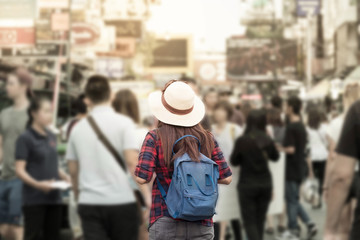 Young Asian woman with hat and backpack traveling in Khaosan Road among people, walking outdoor market in Bangkok, Thailand.
