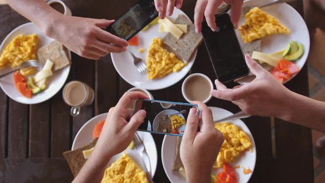 Top view of a group of friends having breakfast in a cafe at wooden table, photographing an omelette, toasts and coffee with mobile phone. slow motion. 4k