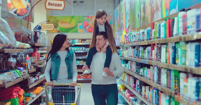 Daughter on shoulders of dad, mom goes next and rolls with trolley in supermarket.